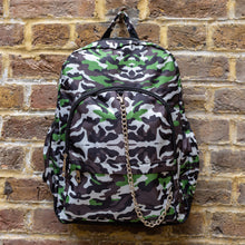 Load image into Gallery viewer, The Savannah Camouflage Backpack hanging outside on a brick wall. The green, brown, khaki, cream and black vegan friendly backpack is facing forward to highlight the two front zip pockets with a silver draping detachable chain, the two side elasticated pockets, the top handle and the main double zip compartment.