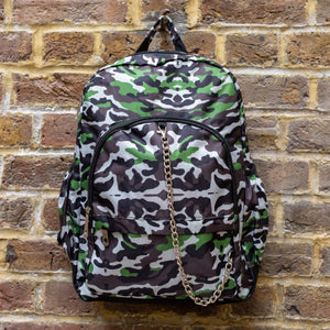 The Savannah Camouflage Backpack hanging outside on a brick wall. The green, brown, khaki, cream and black vegan friendly backpack is facing forward to highlight the two front zip pockets with a silver draping detachable chain, the two side elasticated pockets, the top handle and the main double zip compartment.
