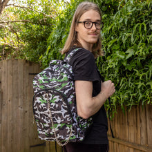 Load image into Gallery viewer, Jack is stood in a garden area modelling the savannah camouflage camo vegan backpack. The bag is facing towards the camera to highlight the front camo print, two front zip pockets, two elastic side pockets, detachable silver chain and top handle.