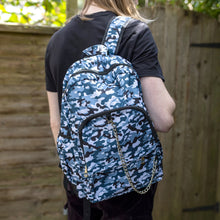 Load image into Gallery viewer, Jack is stood in a garden area modelling the snow camouflage camo vegan backpack. The bag is facing towards the camera to highlight the front camo print, two front zip pockets, two elastic side pockets, detachable silver chain and top handle.
