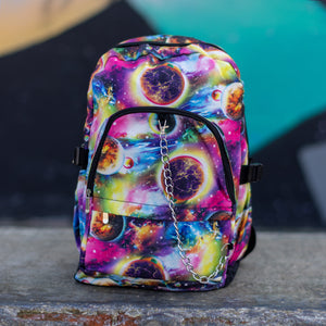 The pink and purple space galaxy vegan backpack sat on a skatepark bench. The backpack is facing forward to highlight the multicoloured space/planet/galaxy print, two zip pockets, two elastic side pockets and detachable silver chain. 