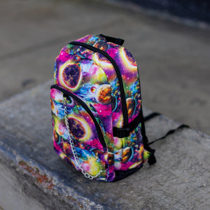 The pink and purple space galaxy vegan backpack sat on a skatepark bench. The backpack is facing forward angled left to highlight the multicoloured space/planet/galaxy print, two zip pockets, two elastic side pockets and detachable silver chain.
