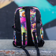 Load image into Gallery viewer, The pink and purple space galaxy vegan backpack sat on a skatepark bench. The backpack is facing away from the camera to highlight the two side pockets, the plain black back and two padded adjustable shoulder straps.
