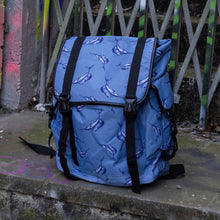Load image into Gallery viewer, The Blue Swordfish Vegan backpack sat near a graffiti urban area. A pastel blue backpack with black buckle and strap detailing with swordfish printed all over. The bag is facing forward to highlight the side buckle pockets, drawstring and buckle close and black piping detailing.