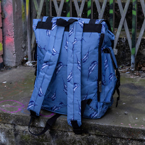 The Blue Swordfish Vegan backpack sat near a graffiti urban area. A pastel blue backpack with black buckle and strap detailing with swordfish printed all over. The bag is facing away from the camera to highlight the side zip pockets, side buckle pockets and adjustable shoulder straps.