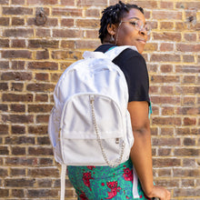 Load image into Gallery viewer, The white backpack with chain is being held by an alternative model's shoulder wearing run and fly dungarees. The vegan friendly bag is facing forward to highlight the top handle, the double zip main compartment, two front zip pockets with a silver decorative detachable draping chain and two elasticated side pockets.