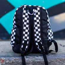 Load image into Gallery viewer, CHOK white checker vegan backpack sat on a skatepark bench in front of a graffiti wall. The bag is facing away to highlight the white and black checkered square printed and adjustable padded shoulder straps, two elastic side pockets and plain back.