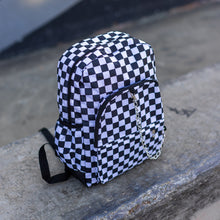Load image into Gallery viewer, CHOK white checker vegan backpack sat on a skatepark bench. The bag is facing forward angled right to highlight the white and black checkered square print, silver detachable chain, two front zip pickets and two side elastic pockets.