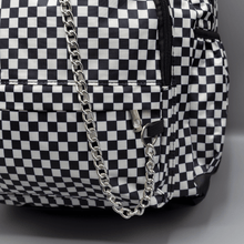 Load image into Gallery viewer, Close up of the White Checkerboard Backpack sat on a grey background. The vegan friendly bag features an all over mini black and white check print with black trim along the silver zips. The bag is facing forward to highlight the two front zip pockets, two side elasticated pockets, the top zip compartment, the top handle and the silver decorative detachable chain draping across the front.
