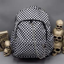 Load image into Gallery viewer, The White Checkerboard Backpack sat on a grey background with a palmistry guide book and two stacked skulls on the left and a phrenology candle and three stacked skulls on the right. The vegan friendly bag features an all over mini black and white check print with black trim along the silver zips. The bag is facing forward to highlight the two front zip pockets, two side elasticated pockets, the top zip compartment, the top handle and the silver decorative detachable chain draping across the front.