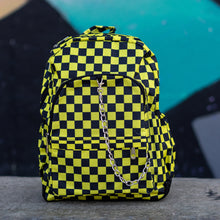 Load image into Gallery viewer, CHOK yellow checker vegan backpack sat on a skatepark bench in front of a graffiti wall. The bag is facing forward to highlight the yellow and black checkered square print, silver detachable chain, two front zip pickets and two side elastic pockets.