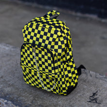 Load image into Gallery viewer, CHOK yellow checker vegan backpack sat on a skatepark bench. The bag is facing forward angled left to highlight the yellow and black checkered square print, silver detachable chain, two front zip pickets and two side elastic pockets.