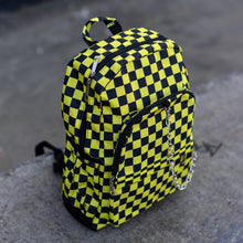 Load image into Gallery viewer, CHOK yellow checker vegan backpack sat on a skatepark bench. The bag is facing forward angled right to highlight the yellow and black checkered square print, silver detachable chain, two front zip pickets and two side elastic pockets.