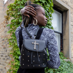 Yolande, an alternative pierced model, wearing a spiderweb long sleeve top with a black pinafore and the GothX Don't Cross Me Tassel Tie Backpack. They are wearing the vegan leather bag on their back with both detachable straps to highlight the cross, skull and hexagonal studs, tassel tie close and the cross chain emblem magnetic clip close flap.