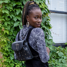 Load image into Gallery viewer, Yolande, an alternative pierced model, wearing a spiderweb long sleeve top with a black pinafore and the GothX Don&#39;t Cross Me Tassel Tie Backpack. They are wearing the vegan leather bag on their back with both detachable straps to highlight the cross, skull and hexagonal studs, tassel tie close and the cross chain emblem magnetic clip close flap.
