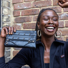 Load image into Gallery viewer, The gothx mini skull head vegan clutch bag being held up by an alternative model with piercings sat outside in front of a brick wall wearing a run and fly black boilersuit whilst laughing. The vegan leather clutch makeup bag is facing forward to show two lines of stitching detailing and metal skull studs with two lower faux zip decorations.
