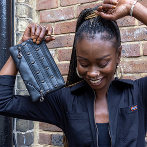 The gothx mini skull head vegan clutch bag being held up by an alternative model with piercings sat outside in front of a brick wall wearing a run and fly black boilersuit whilst laughing. The vegan leather clutch makeup bag is facing forward to show two lines of stitching detailing and metal skull studs with two lower faux zip decorations.