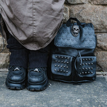 Load image into Gallery viewer, The GothX twin pocket skull vegan backpack sat outside next to a brown brick wall next to a y2k grunge styled model wearing a cargo skirt, leg warmers and black buffalo platform trainers. The vegan leather gothic style bag is facing forward to highlight the diamante effect skull, skull embossed vegan leather front flap, tassel tie cords and two silver studded front pockets.
