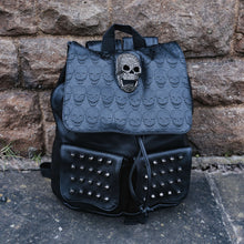 Load image into Gallery viewer, The GothX twin pocket skull vegan backpack sat outside next to a brown brick wall. The vegan leather gothic style bag is facing forward to highlight the diamante effect skull, skull embossed vegan leather front flap, tassel tie cords and two silver studded front pockets.
