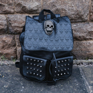 The GothX twin pocket skull vegan backpack sat outside next to a brown brick wall. The vegan leather gothic style bag is facing forward to highlight the diamante effect skull, skull embossed vegan leather front flap, tassel tie cords and two silver studded front pockets.