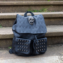 Load image into Gallery viewer, The GothX twin pocket skull vegan backpack sat on a concrete stairway outside. The vegan leather gothic style bag is facing forward to highlight the diamante effect skull, skull embossed vegan leather front flap, tassel tie cords and two silver studded front pockets.

