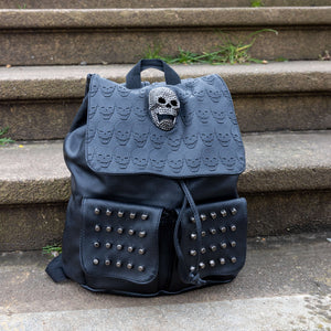 The GothX twin pocket skull vegan backpack sat on a concrete stairway outside. The vegan leather gothic style bag is facing forward to highlight the diamante effect skull, skull embossed vegan leather front flap, tassel tie cords and two silver studded front pockets.