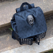 Load image into Gallery viewer, The GothX twin pocket skull vegan backpack sat on a concrete stairway outside. The vegan leather gothic style bag is facing forward to highlight the diamante effect skull, skull embossed vegan leather front flap, tassel tie cords and two silver studded front pockets.
