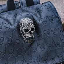 Load image into Gallery viewer, Close up of the GothX twin pocket skull vegan backpack sat outside next to a brown brick wall. The vegan leather gothic style bag is facing forward to highlight the diamante effect skull, skull embossed vegan leather front flap, tassel tie cords and two silver studded front pockets.
