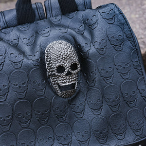 Close up of the GothX twin pocket skull vegan backpack sat outside next to a brown brick wall. The vegan leather gothic style bag is facing forward to highlight the diamante effect skull, skull embossed vegan leather front flap, tassel tie cords and two silver studded front pockets.