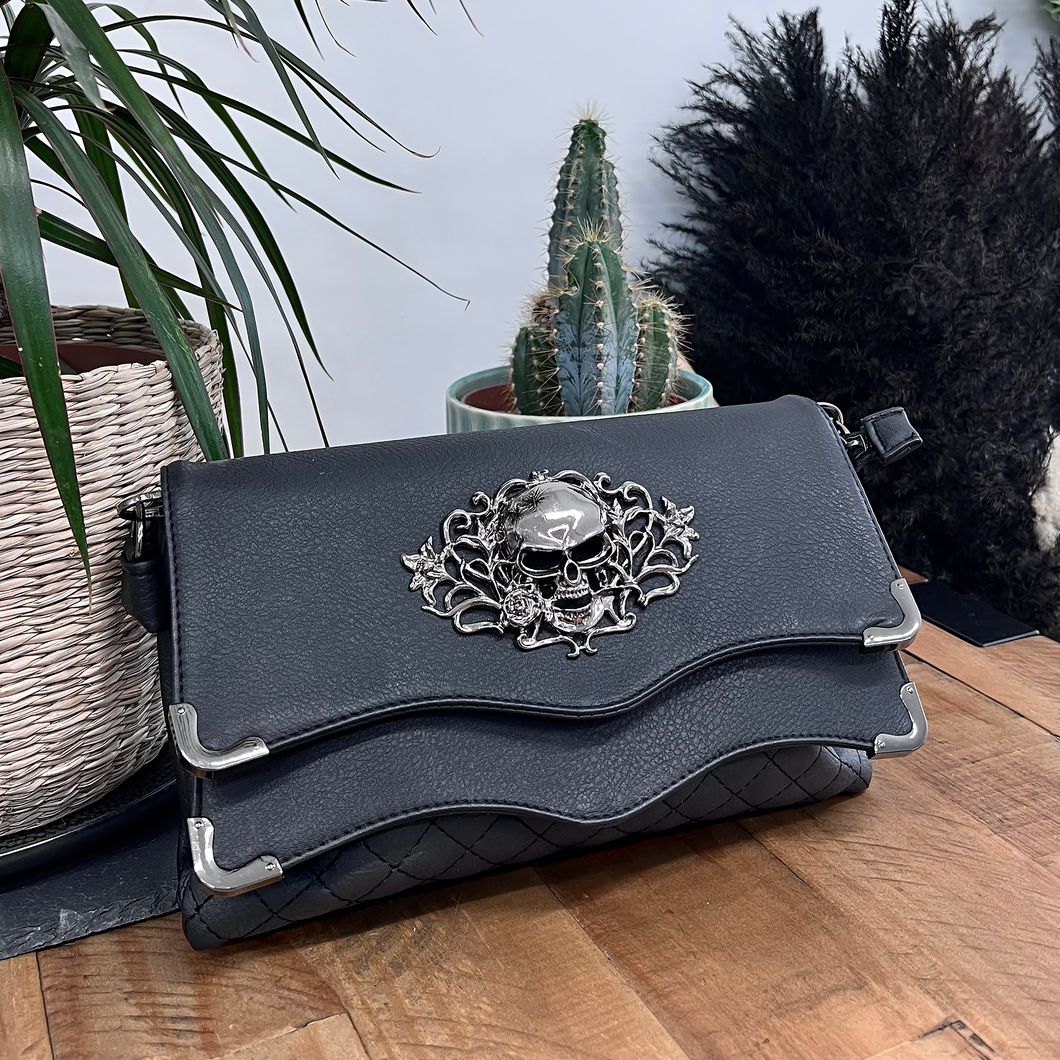 The GothX Skulls and Roses Quilted Clutch Bag sat on a wooden table with black and green plant foliage behind the bag. The vegan leather clutch bag is facing forward to highlight the two magnetic clip close flaps with metal corners, a stitch quilted front, a skulls and roses metal centrepiece and two D rings on either side for a detachable strap. The mini bag is inspired by gothic grunge witchy fashion.
