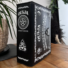 Load image into Gallery viewer, The GothX Ouija Spirit Book Mini Bag sat on a wooden table with green foliage and plants behind. The bag is facing sideways to highlight the ouija spirit book white printed detailing on the black book bag with the 3D planchette stitching. Bag is inspired by witchy style and necromancy.
