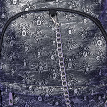 Load image into Gallery viewer, A close up of the Rustic Silver Spider Backpack sat on a purple background. The bag is facing forward to highlight the two zip front pockets, two elasticated side pockets, the double zip main compartment with a silver draping chain across the front. The vegan friendly faux leather bag has 3d embossed spiders in varying sizes with brushed black and silver tones all over.

