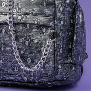 A close up of the Rustic Silver Spider Backpack sat on a purple background. The bag is facing forward to highlight the two zip front pockets, two elasticated side pockets, the double zip main compartment with a silver draping chain across the front. The vegan friendly faux leather bag has 3d embossed spiders in varying sizes with brushed black and silver tones all over.