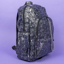 Load image into Gallery viewer, The Rustic Silver Spider Backpack sat on a purple background. The bag is facing right to highlight the two zip front pockets, two elasticated side pockets, the double zip main compartment with a silver draping chain across the front. The vegan friendly faux leather bag has 3d embossed spiders in varying sizes with brushed black and silver tones all over.

