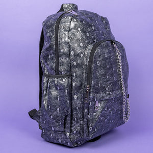 The Rustic Silver Spider Backpack sat on a purple background. The bag is facing right to highlight the two zip front pockets, two elasticated side pockets, the double zip main compartment with a silver draping chain across the front. The vegan friendly faux leather bag has 3d embossed spiders in varying sizes with brushed black and silver tones all over.