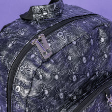 Load image into Gallery viewer, A close up of The Rustic Silver Spider Backpack sat on a purple background. The bag is facing right to highlight the two zip front pockets, two elasticated side pockets, the double zip main compartment, top handle with a silver draping chain across the front. The vegan friendly faux leather bag has 3d embossed spiders in varying sizes with brushed black and silver tones all over.

