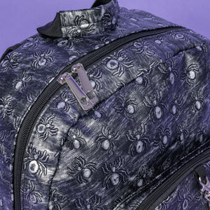A close up of The Rustic Silver Spider Backpack sat on a purple background. The bag is facing right to highlight the two zip front pockets, two elasticated side pockets, the double zip main compartment, top handle with a silver draping chain across the front. The vegan friendly faux leather bag has 3d embossed spiders in varying sizes with brushed black and silver tones all over.