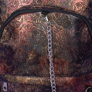 Close up of the The Rustic Skulls & Roses Backpack sat on a purple background. The bag is facing forward to highlight the two zip front pockets, two elasticated side pockets, the double zip main compartment with a silver draping chain across the front. The bag is varying colours of bronze, gold, rose and brown with black 3d embossed sugar skulls, roses and swirls all over.