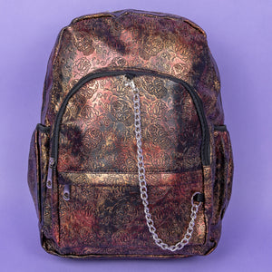 The Rustic Skulls & Roses Backpack sat on a purple background. The bag is facing forward to highlight the two zip front pockets, two elasticated side pockets, the double zip main compartment with a silver draping chain across the front. The bag is varying colours of bronze, gold, rose and brown with black 3d embossed sugar skulls, roses and swirls all over.