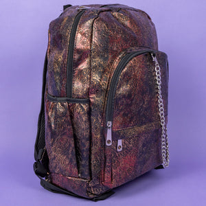 The Rustic Skulls & Roses Backpack sat on a purple background. The bag is facing forward angled right to highlight the two zip front pockets, two elasticated side pockets, the double zip main compartment with a silver draping chain across the front. The bag is varying colours of bronze, gold, rose and brown with black 3d embossed sugar skulls, roses and swirls all over.