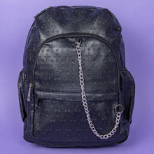Load image into Gallery viewer, The Black Embossed Cross Backpack sat on a purple background. The backpack is facing forward to highlight the two front silver zip pockets, two elasticated side pockets, main double zip compartment and a silver decorative detachable silver chain draping across the front. The bag is made of vegan friendly leather with 3d embossed crosses in varying sizes all over.
