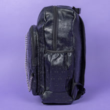 Load image into Gallery viewer, The Black Embossed Cross Backpack sat on a purple background. The backpack is facing left to highlight the two front silver zip pockets, two elasticated side pockets, main double zip compartment and a silver decorative detachable silver chain draping across the front. The bag is made of vegan friendly leather with 3d embossed crosses in varying sizes all over.
