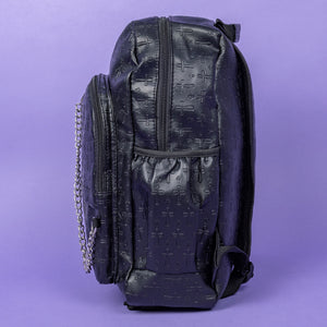 The Black Embossed Cross Backpack sat on a purple background. The backpack is facing left to highlight the two front silver zip pockets, two elasticated side pockets, main double zip compartment and a silver decorative detachable silver chain draping across the front. The bag is made of vegan friendly leather with 3d embossed crosses in varying sizes all over.