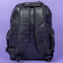 Load image into Gallery viewer, The Black Embossed Cross Backpack sat on a purple background. The backpack is facing away from the camera to highlight the two elasticated side pockets, padded shoulder straps and top handle. The bag is made of vegan friendly leather with 3d embossed crosses in varying sizes all over.
