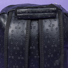 Load image into Gallery viewer, A close up of the Black Embossed Cross Backpack sat on a purple background. The backpack is facing away from the camera to highlight the two elasticated side pockets, padded shoulder straps and top handle. The bag is made of vegan friendly leather with 3d embossed crosses in varying sizes all over.
