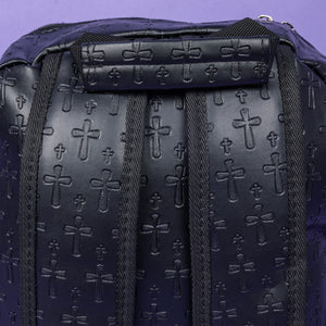 A close up of the Black Embossed Cross Backpack sat on a purple background. The backpack is facing away from the camera to highlight the two elasticated side pockets, padded shoulder straps and top handle. The bag is made of vegan friendly leather with 3d embossed crosses in varying sizes all over.