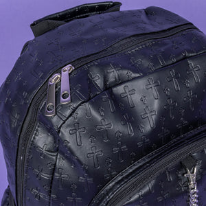 A close up of the Black Embossed Cross Backpack sat on a purple background. The backpack is facing forward to highlight the two front silver zip pockets, two elasticated side pockets, main double zip compartment and a silver decorative detachable silver chain draping across the front. The bag is made of vegan friendly leather with 3d embossed crosses in varying sizes all over.