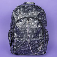 Load image into Gallery viewer, The Rustic Skulls Backpack sat on a purple background. The bag is facing forward to highlight the two front zip pockets, two elasticated side pockets, main zip compartment, the top handle and the detachable decorative silver chain. All over the backpack is an embossed 3d texture skulls and skull and crossbones on a faux leather material in a brushed black and silver grunge style.
