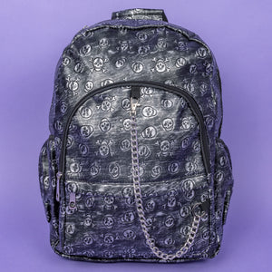 The Rustic Skulls Backpack sat on a purple background. The bag is facing forward to highlight the two front zip pockets, two elasticated side pockets, main zip compartment, the top handle and the detachable decorative silver chain. All over the backpack is an embossed 3d texture skulls and skull and crossbones on a faux leather material in a brushed black and silver grunge style.