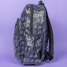 Load image into Gallery viewer, The Rustic Skulls Backpack sat on a purple background. The bag is facing left to highlight the two front zip pockets, two elasticated side pockets, main zip compartment, the top handle and the detachable decorative silver chain. All over the backpack is an embossed 3d texture skulls and skull and crossbones on a faux leather material in a brushed black and silver grunge style.
